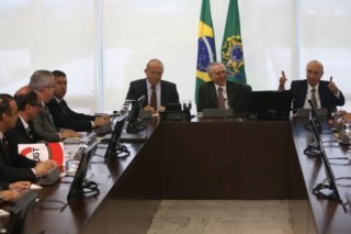 Sete homens reunidos ao redor de uma mesa. Atrás aparece a bandeira do Brasil.