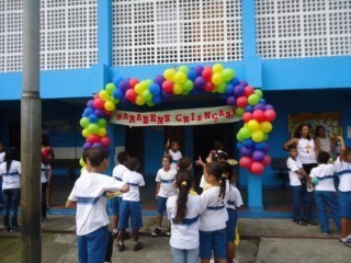 Crianças com uniformes escolares no pátio. Na porta de entrada uma faixa com balões coloridos, onde diz 