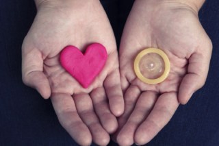Sobre a palma da mão direita tem um coração feito de massa de modelar e sobre a esquerda um preservativo masculino.