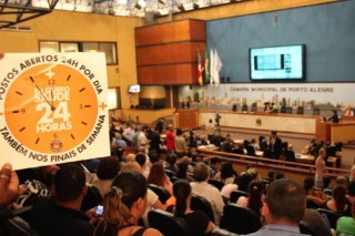 Plenário da câmara de vereadores. A esquerda da foto aparece um cartaz com uma figura de relógio onde diz: eu quero postos de saúde 24 horas.