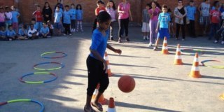 Menina brincando com uma bola num pátio de escola. ao fundo várias crianças olhando.