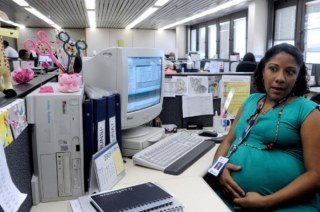 Mulher grávida sentada em um escritório. Sob a mesa computador e papéis.