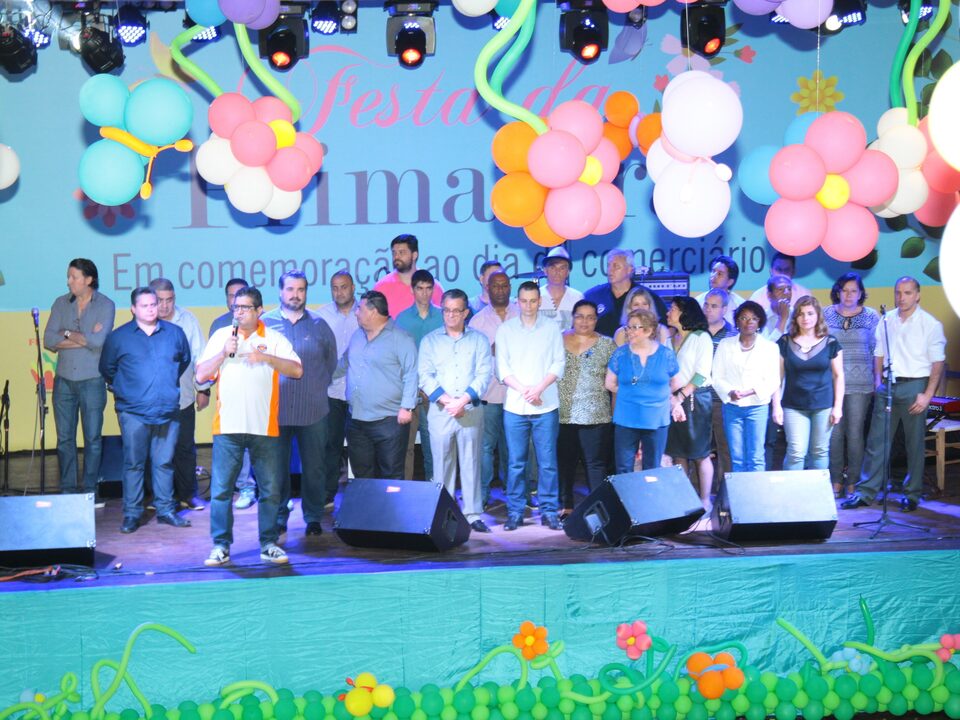 Membros da diretoria do sindicato no palco do Centro de Eventos Casa do Gaúcho. Presidente Nilton Neco discursa. Na parte superior o palco está decorado com balões em formato de flores.