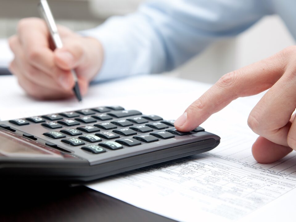 Mão direita, que indica ser masculina, segura uma caneta. A mão esquerda mexe em uma calculadora que está sob uma mesa de escritório.