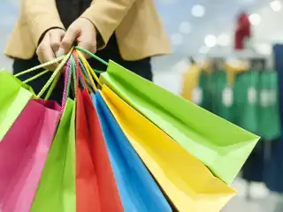 Mãos segurando sete sacolas coloridas de compras.Ao fundo, desfocado, aparece uma arara de roupas.