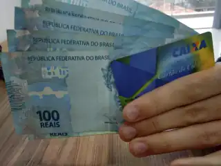 Mão segurando notas de cem reais e um cartão da Caixa Econômica Federal