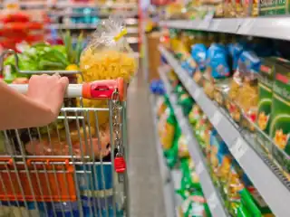 Mão empurrando carrinho de supermercado cheio de alimentos. Ao lado prateleiras com pacotes de macarrão.