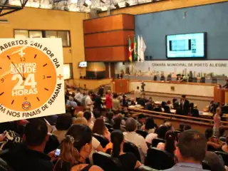 Plenário da câmara de vereadores. A esquerda da foto aparece um cartaz com uma figura de relógio onde diz: eu quero postos de saúde 24 horas.