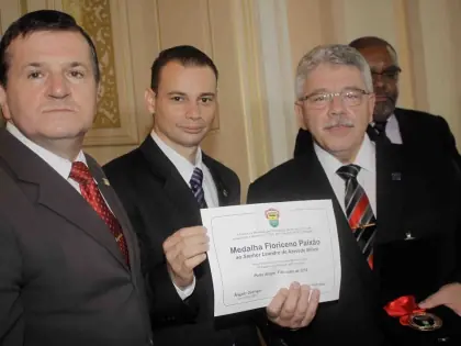 Três homens. O do centro segura um certificado e o da lateral direita uma medalha.