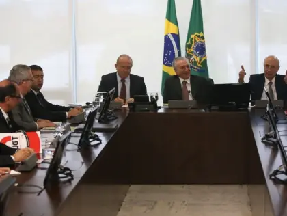 Sete homens reunidos ao redor de uma mesa. Atrás aparece a bandeira do Brasil.