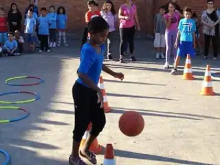 Menina brincando com uma bola num pátio de escola. ao fundo várias crianças olhando.