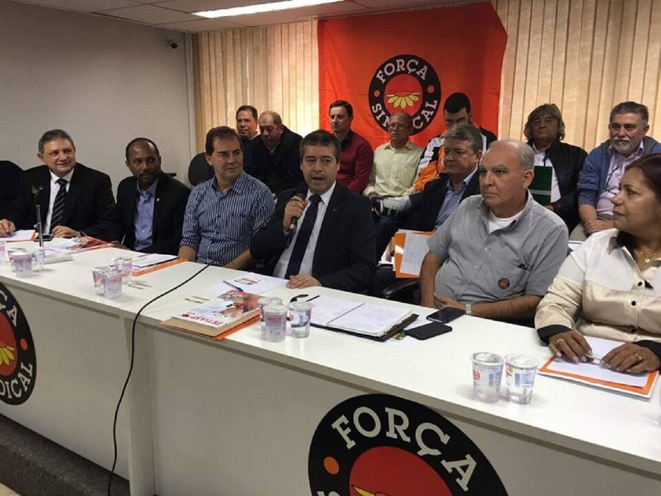 Pessoas sentadas em uma bancada. Atrás bandeira da força Sindical