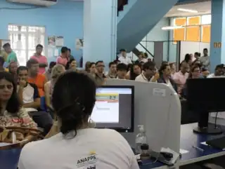 Atendente sentada de costas mexendo no computador. Pessoas sentadas esperando atendimento.