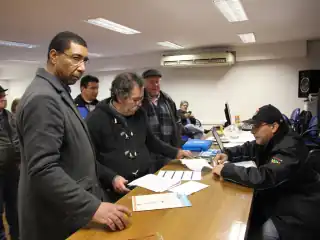 Homens em pé conferindo papéis. Um está sentado.