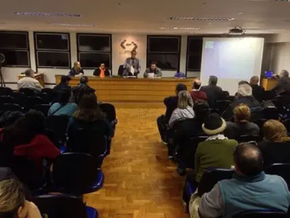 Pessoas em um auditório. Ao fundo uma bancada com pessoas sentadas.