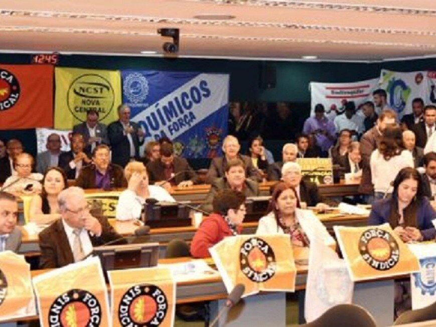 Pessoas sentadas num plenário. Em cada mesa bandeiras com a logomarca da Força Sindical.