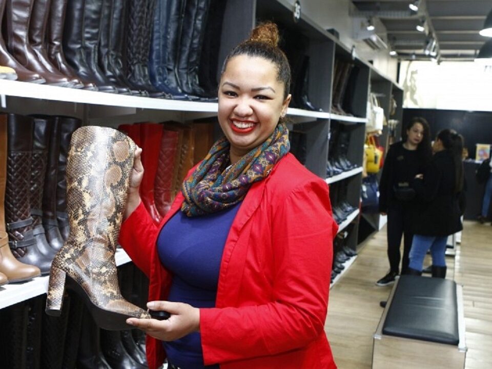 Mulher segurando um pé de bota. Ao lado uma prateleira com vários pares de botas.