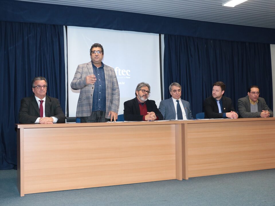 Seis homens sentado atrás de um balcão num palco. O segundo, da esquerda para a direita está em pé falando.