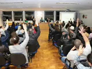 Pessoas sentadas em um auditório com o braço direito levantado.