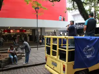 Dois homens em cima de um carro de som. Um fala no microfone. Ao fundo lojas Renner.