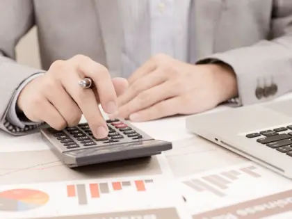 Homem usando calculadora. Sob mesa também há um notebook.