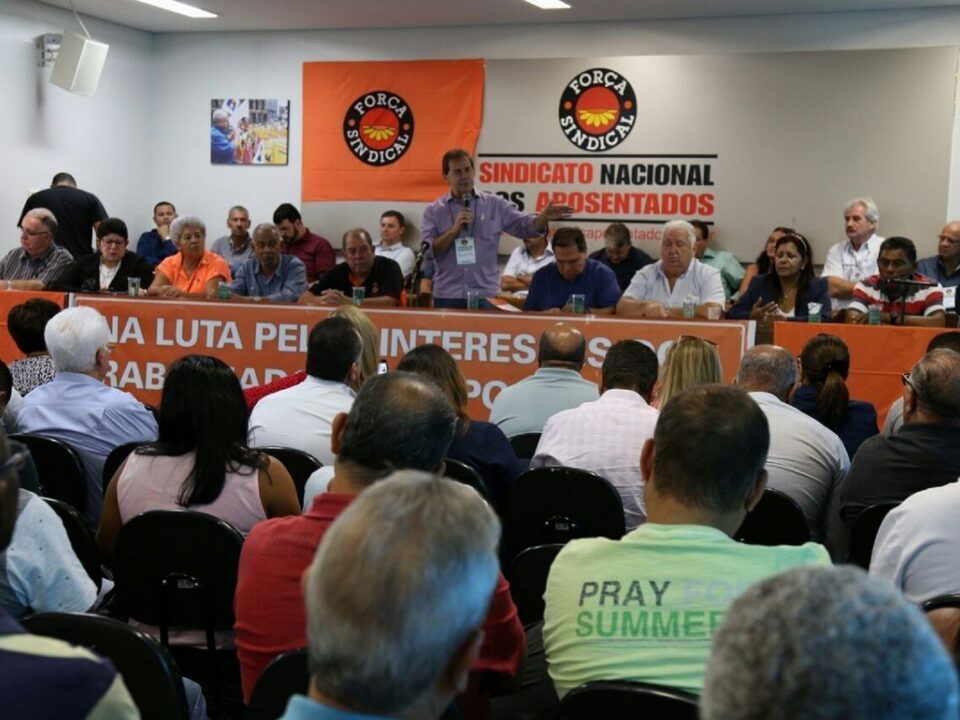 Pessoas sentadas em um auditório. Homem em pé falando no microfone