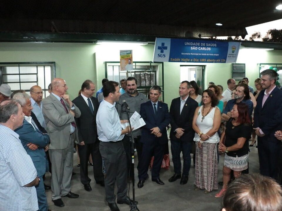 Pessoas em pé. Homem discursando no microfone.