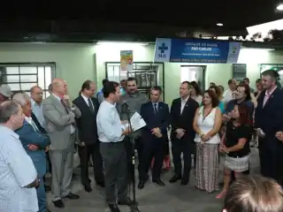 Pessoas em pé. Homem discursando no microfone.