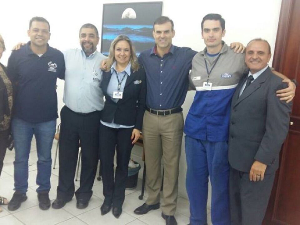 Sete pessoas em pé , numa sala, posando pra foto. Duas mulheres e cinco homens