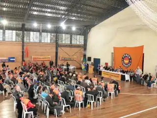 pessoas sentadas em cadeiras na quadra de um ginásio. Bandeira da Força Sindical ao fundo.