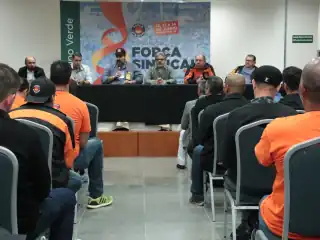 Pessoas sentadas em um auditório. Na mesa ao fundo , faixa da força sindical.