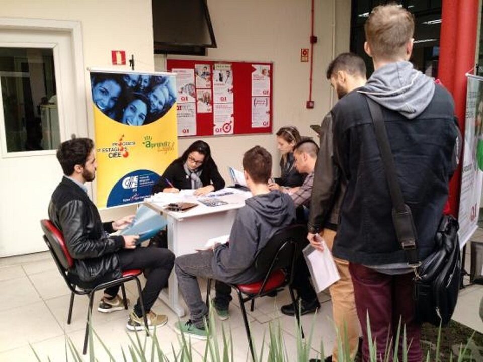 Jovens sentados em uma mesa. Menina escrevendo. Dois meninos em pé.