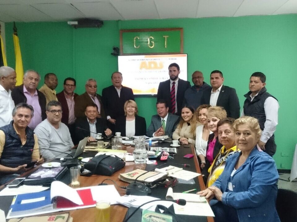 Grupo de pessoas, homens e mulheres, alguns sentados em torno de uma mesa, outros em pé ao fundo da sala. Parede verde com bandeiras ao lado esquerdo e uma televisão no centro.