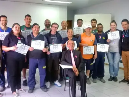 Pessoas em pé segurando um certificado na mão. Um homem em frente a eles sentado numa cadeira.